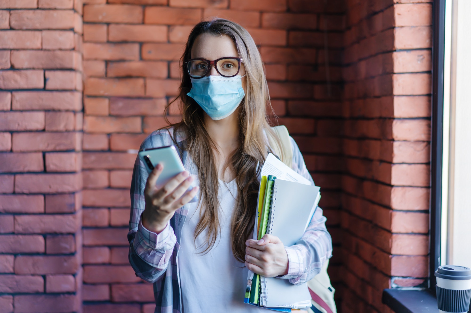female student in protective medical mask with phone and textbooks in college.
