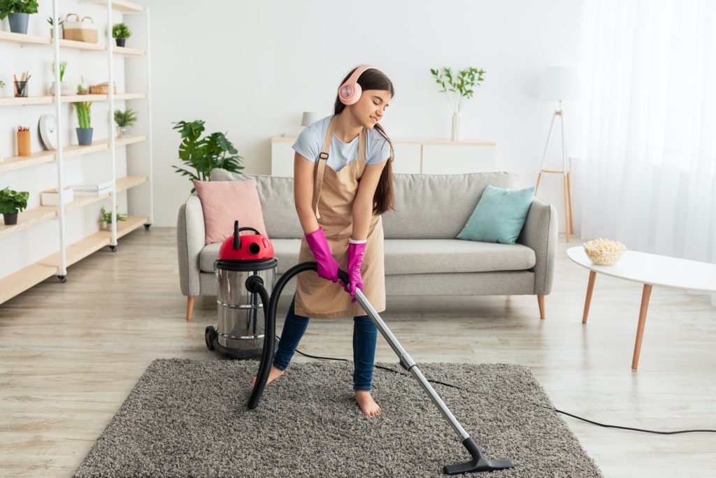 Happy Indian teen girl cleaning her home, wearing headphones, listening to music while vacuuming