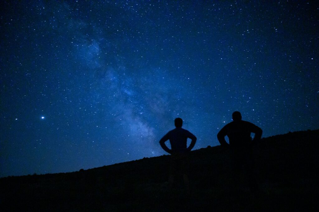 Magical view of silhouettes of two men stargazing, admiring the Milky way and the blue night sky