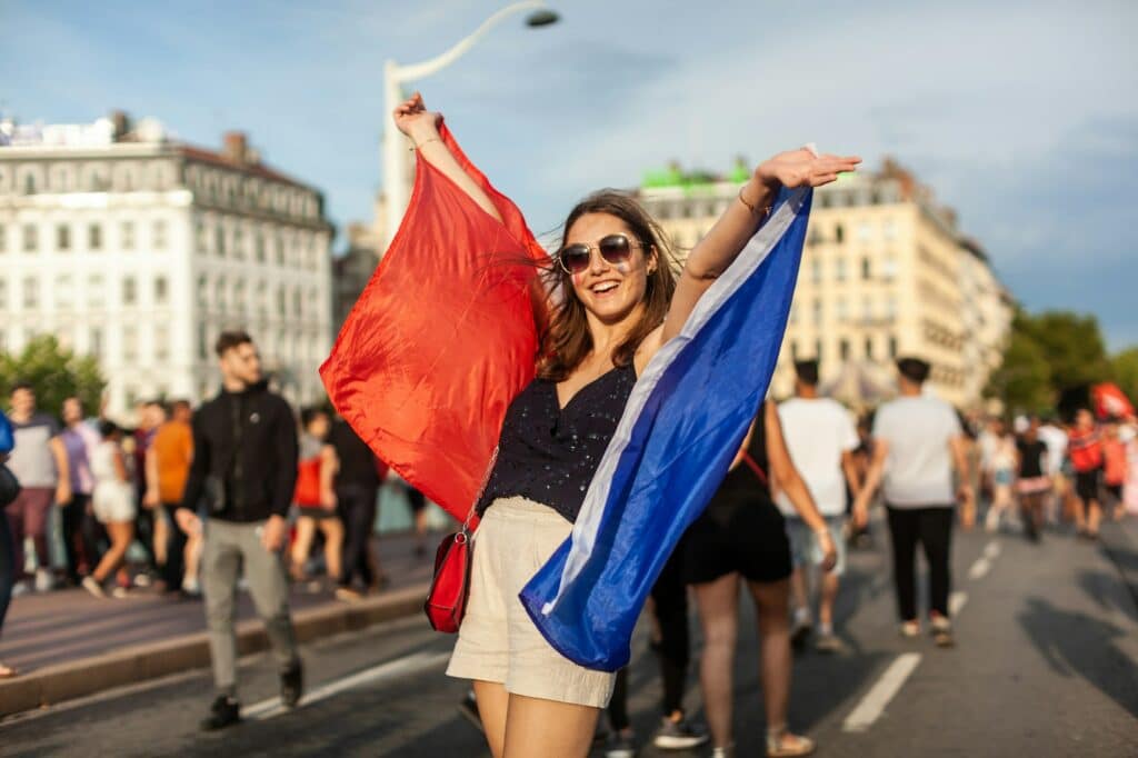 Young French woman raising her arms in the air from excitement carrying a French flag wearing shorts