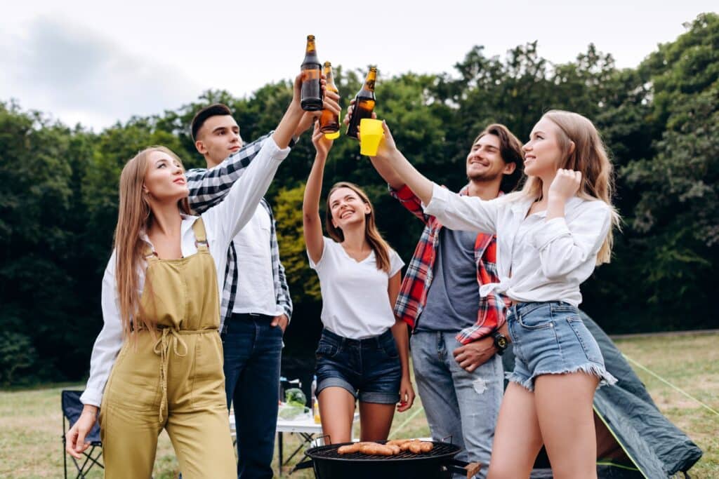 A company of friends raising a bottles with a beer in the camping . - Image