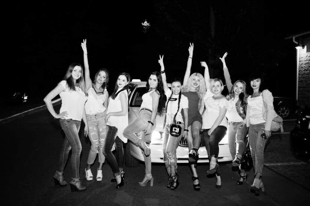 Cool bride in red dress posing with her bridesmaids against the limousin at bachelorette party.